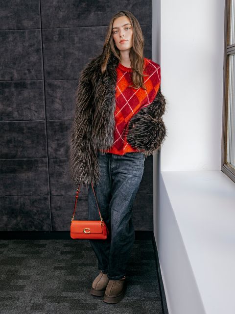 Eine Frau in einem roten Argyle-Pullover, einem Kunstpelzmantel, grauen Jeans und hellbraunen Schuhen lehnt an einer Wand in der Nähe eines Fensters und hält eine rote Handtasche in der Hand.