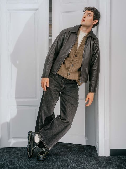 Ein Mann in einer dunklen Hose, einer braunen Jacke und einem Ledermantel lehnt an einer weißen Tür und blickt mit überraschtem Gesichtsausdruck nach oben.