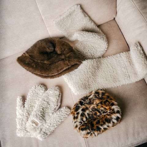 Auf einem beigen Sofa liegen vier winterliche Accessoires aus Pelz, darunter zwei Mützen, ein Schal und Handschuhe.
