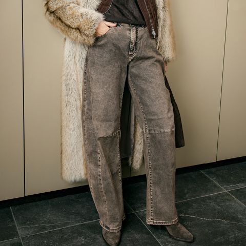 Eine Person, die eine braune Jeans mit weitem Bein, einen Pelzmantel und spitze Stiefel trägt und eine Tasche in der Hand hält, steht vor beigen Schränken.