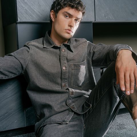 Ein Mann mit kurzen Locken, der ein graues Jeanshemd und Jeans trägt, sitzt auf dem Boden, lehnt an einer dunklen, modernen Wand und blickt in die Kamera.