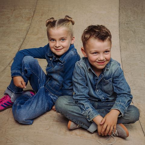Zwei Kinder in Jeansoutfits sitzen auf einem Holzboden und lächeln in die Kamera. Das eine trägt rosa Turnschuhe, das andere hellbraune Schuhe.
