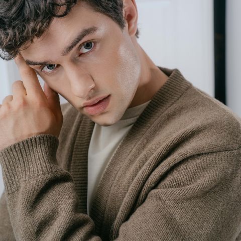 Ein junger Mann in einem beigen Pullover, der mit der Hand an der Schläfe posiert.