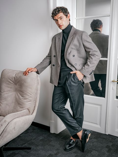 Ein Mann, der einen grauen karierten Blazer, einen schwarzen Rollkragenpullover und eine dunkle Hose trägt, lehnt an einem Stuhl.