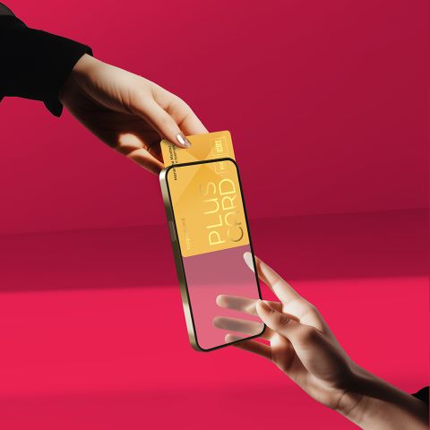 Zwei Hände halten eine goldene Plus-Karte über ein transparentes Smartphone vor einem rosa Hintergrund.