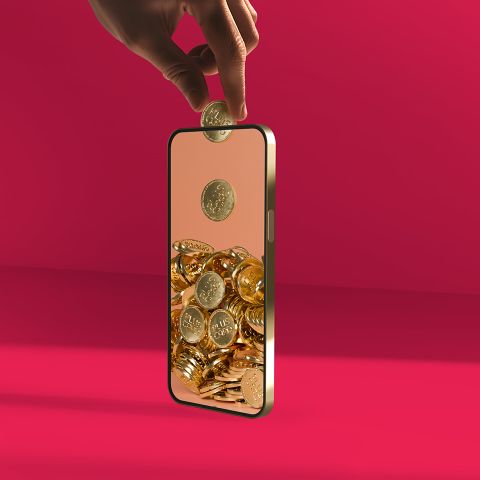Eine Hand lässt eine Goldmünze in ein Smartphone fallen, in dem sich bereits ein Haufen Goldmünzen ansammelt.