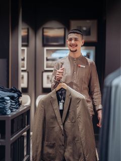 Eine lächelnde Verkäuferin in einem beigen Leinenhemd hält in einem Bekleidungsgeschäft einen braunen Blazer hoch.