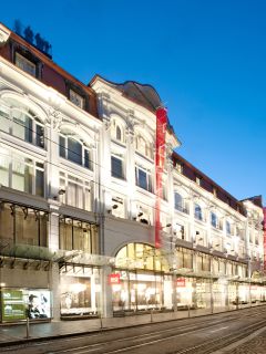 Die kunstvoll verzierte Fassade des KÖ-Einkaufszentrums in der Abenddämmerung, mit Straßenbahnschienen im Vordergrund.
