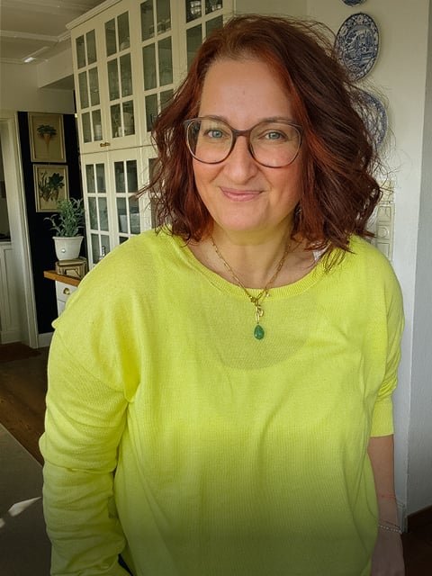 Eine Frau mit rotbraunen Haaren und Brille, die einen leuchtend gelben Pullover und eine Halskette mit Anhänger trägt.