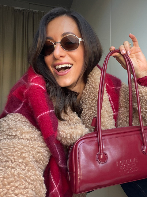 Smiling woman with dark hair wearing round sunglasses, a tan teddy coat and red scarf, holding a red handbag by its handles.