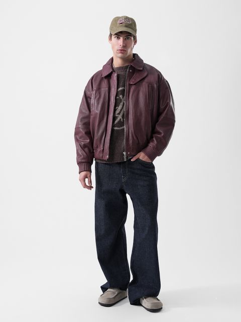 Ein Mann, der eine burgunderrote Leder-Bomberjacke, einen braunen gemusterten Pullover, dunkelblaue Jeans, eine Baseballkappe und Slipper trägt.