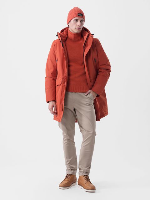Ein Mann, der eine orangefarbene Mütze, einen Rollkragenpullover und einen Parka trägt, dazu eine beige Hose und braune Lederstiefel.