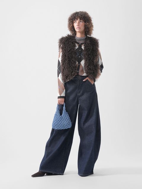 Ein Model trägt eine braune Kunstfellweste über einer Strickjacke mit Argyle-Muster sowie eine dunkelblaue Jeans mit weitem Bein und hält eine kleine blaue Handtasche mit Strukturmuster in der Hand.